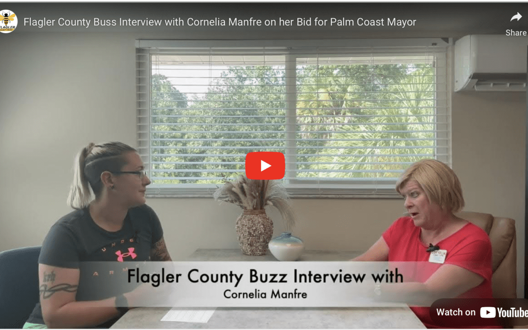 Two individuals are sitting at a table with a centerpiece in a room with a large window Text overlay reads, "Flagler County Buzz Interview with Cornelia Manfre on her Bid for Palm Coast Mayor " A red play button indicates a video